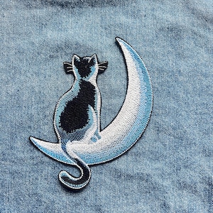 Op de afbeelding: Een geborduurde patch met een zwarte kat die op een halve maan zit. De kat heeft blauwe accenten en is van achteren te zien. De halve maan is in wit en lichtblauw geschaduwd, met een zwarte rand die het ontwerp omlijnt.