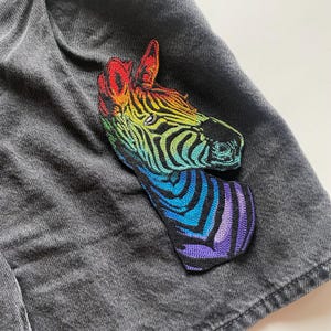 Op de afbeelding: Een regenboogkleurige zebrakop-patch, geborduurd met een gedetailleerd ontwerp. De patch is genaaid op een donkergrijze denim stof.
