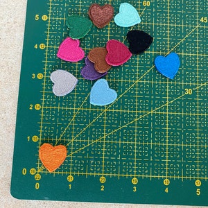 May include: Assortment of felt heart patches in various colors, including red, pink, blue, green, brown, and gray. The patches are arranged on a green cutting mat with a grid pattern.