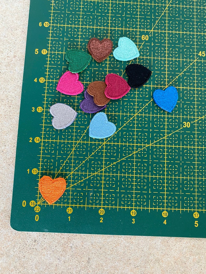 May include: Assortment of felt heart patches in various colors, including red, pink, blue, green, brown, and gray. The patches are arranged on a green cutting mat with a grid pattern.