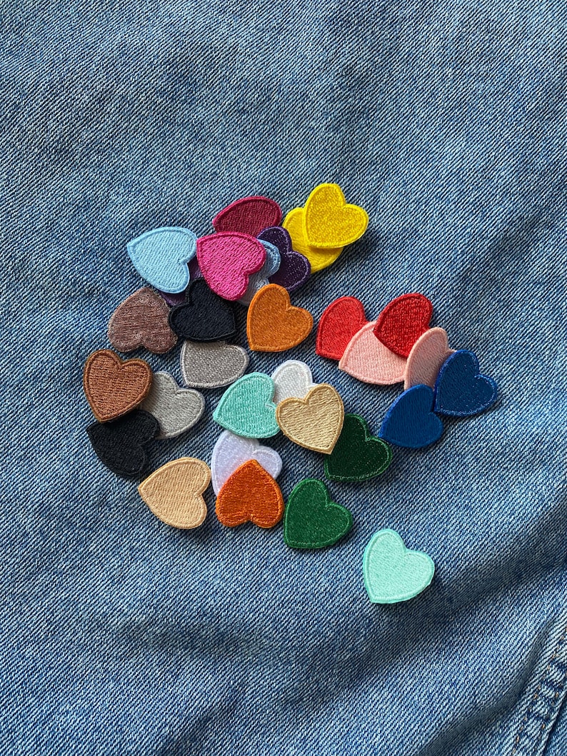 May include: A collection of colorful felt heart patches in various shades of red, pink, blue, green, yellow, orange, brown, and white. The patches are arranged in a random pattern on a blue denim background.