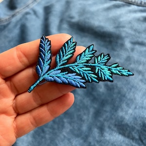 May include: Embroidered leaf patch in shades of blue and teal, with intricate stitching details. The patch features a black outline and is designed to be sewn onto fabric. This decorative patch adds a touch of nature-inspired flair to clothing or accessories.