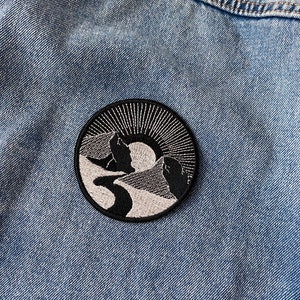 May include: A black and white embroidered patch featuring a mountain range and a sun in the background. The patch is circular and has a white outline.