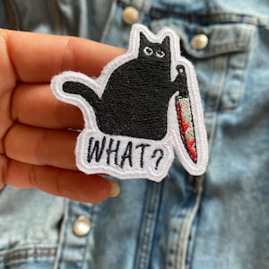 Peut inclure: Un chat noir tenant un couteau ensanglanté avec le texte "WHAT?" brodé sur un patch blanc.
