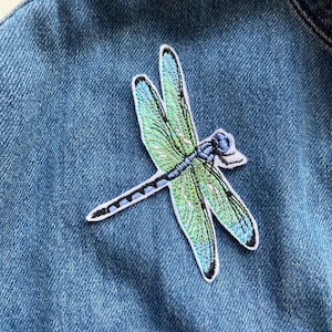 Peut inclure: Un patch brodé représentant une libellule bleue et verte. La libellule a un design détaillé avec des ailes irisées.