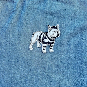 Può includere: Una toppa in bianco e nero raffigurante un bulldog francese con una maglietta a righe. La toppa è su uno sfondo di denim blu. Il cane è in piedi, rivolto verso destra, con linee dettagliate.