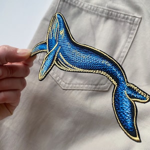Può includere: Una toppa ricamata a forma di balena blu e dorata. La balena è rivolta a destra e presenta un design dettagliato.