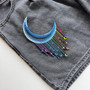 Może przedstawiać: Haftowana naszywka przedstawiająca niebieski półksiężyc z tęczowymi akcentami gwiazd i księżyców. Naszywka jest przyszyta do szarych dżinsowych szortów. Naszywka ma motyw niebieski.