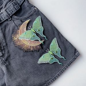 Könnte beinhalten: Zwei grüne und weiße gestickte Motten-Patches auf einer dunkelgrauen Jeansjacke. Die Motten sind in einer Halbmondform mit einem Sonnenstrahlen-Design.