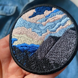Peut inclure: Patch brodé représentant une scène de paysage avec des nuages bleus, roses et blancs au-dessus d'une chaîne de montagnes bleu foncé et noire.