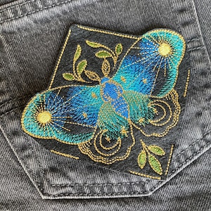 May include: A blue and green embroidered patch of a moth with gold thread details on a black background. The moth has a stylized design with a crescent moon and stars.
