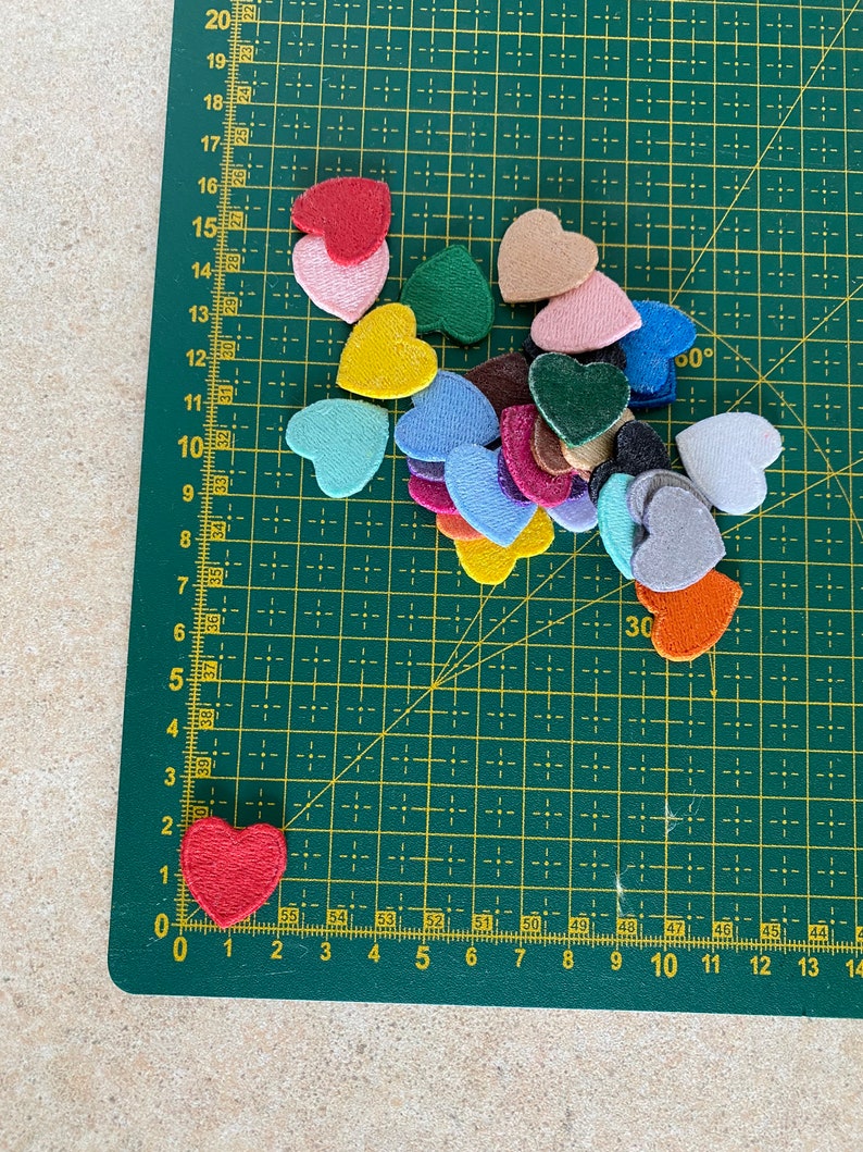 May include: A variety of felt hearts in different colors, including red, pink, yellow, blue, green, and brown, are arranged on a green cutting mat. The hearts are all different sizes and shapes.