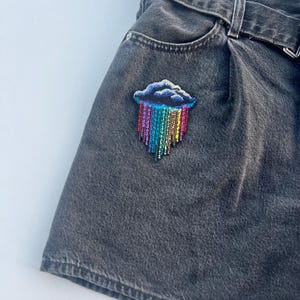 May include: A colorful embroidered patch of a cloud with a rainbow rain design. The patch is sewn onto a pair of dark gray denim shorts. The shorts have a belt and a pocket.