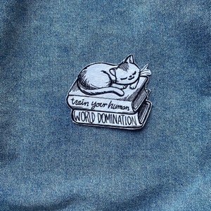 Puede incluir: Un pin en blanco y negro con un gato durmiendo sobre dos libros. Los libros tienen el texto "train your human" y "WORLD DOMINATION". El pin está sobre un fondo de mezclilla azul.