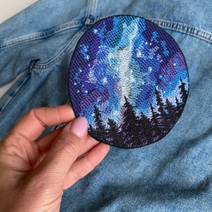 May include: A circular embroidered patch depicting a night sky scene with a forest outline. The patch uses blue, purple, and white threads. It is shown on a denim jacket.