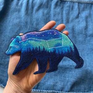 May include: An iron-on patch featuring a blue, green and purple embroidered bear with a starry sky design.