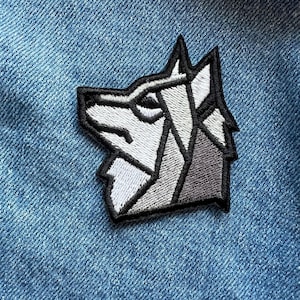 May include: Embroidered patch featuring a geometric wolf head design in shades of gray and white, outlined in black. The patch is sewn onto a blue denim fabric, creating a contrast in textures and colors. This patch is suitable for jackets, bags, or other fabric items.