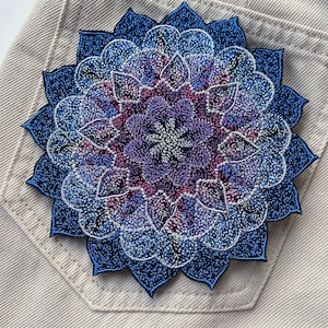 Peut inclure: Un patch brodé de mandala bleu et violet avec un motif en fil argenté. Le patch est sur un fond de tissu brun clair.