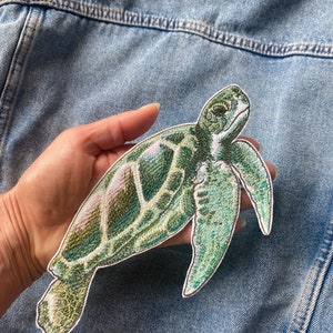Può includere: Toppa ricamata di tartaruga marina verde e bianca. La tartaruga è rivolta a destra e ha un guscio e pinne dettagliate.
