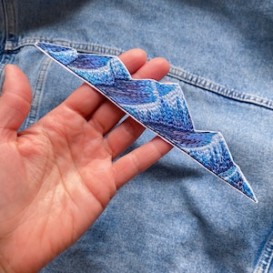 Peut inclure: Un patch brodé bleu et blanc en forme de chaîne de montagnes. Le patch est fait de tissu et a une surface texturée.