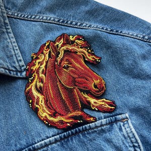 Puede incluir: Un parche bordado que muestra la cabeza de un caballo con una crin fluida en tonos rojo oscuro, naranja y amarillo, que se asemejan a llamas. El parche está sujeto a una chaqueta de mezclilla azul con costuras visibles y un bolsillo.