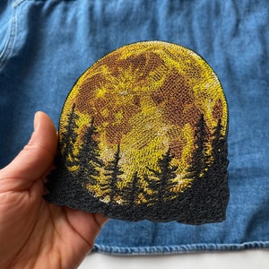 Könnte beinhalten: Gestickter Aufnäher mit einem goldenen Mond und silhouettierten Bäumen. Der Aufnäher hat ein detailliertes Design mit Gold-, Braun- und Schwarztönen. Dieser dekorative Aufnäher eignet sich zum Aufnähen auf Kleidung oder Accessoires.