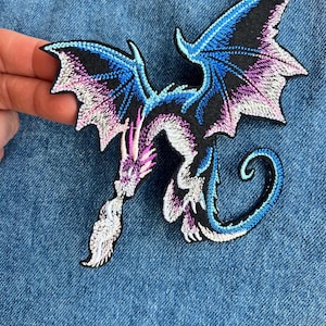 Peut inclure: Écusson brodé en forme de dragon noir, avec des ailes bleues et violettes, et une flamme blanche. Le dragon est finement détaillé avec une queue incurvée, sur fond de denim bleu.
