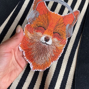 Könnte beinhalten: Gestickter Aufnäher eines Fuchses mit Kopfhörern. Der Fuchs ist orange mit braunem Fell und hat die Augen geschlossen. Die Kopfhörer sind grau.