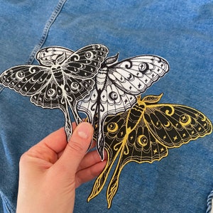 Peut inclure: Trois patchs thermocollants brodés en noir et blanc de papillons de nuit avec des motifs de lune et d'étoiles. Les papillons de nuit sont dans différentes poses avec leurs ailes déployées.