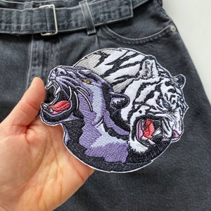 Puede incluir: Parche bordado con un pantera negra y un tigre blanco enfrentados con la boca abierta. El parche es circular y tiene un contorno blanco.
