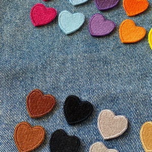May include: Assortment of felt heart patches in various colors, including pink, purple, orange, brown, black, and beige. The patches are arranged on a blue denim background.