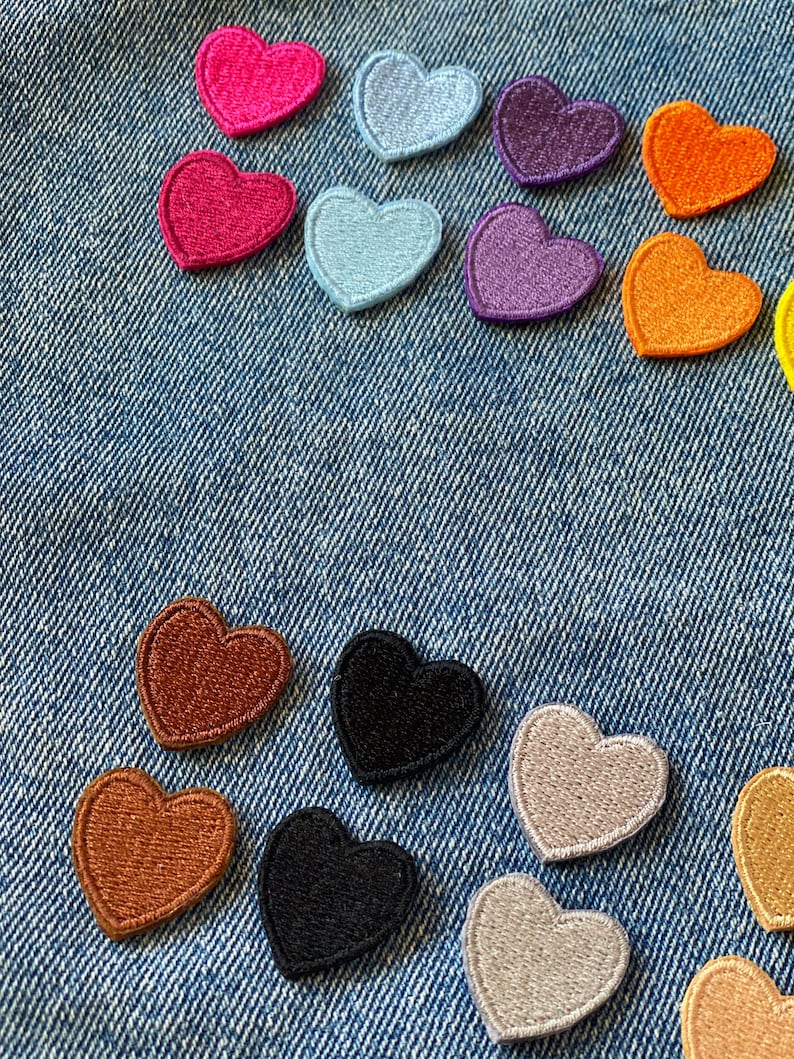 May include: Assortment of felt heart patches in various colors, including pink, purple, orange, brown, black, and beige. The patches are arranged on a blue denim background.