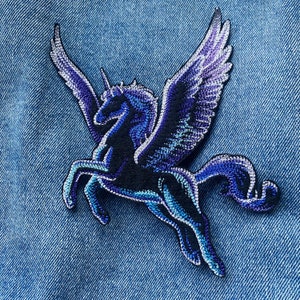 Könnte beinhalten: Ein gestickter Aufnäher mit einem geflügelten Einhorn mit einem silbernen Horn. Das Einhorn befindet sich in einer laufenden Pose mit ausgebreiteten Flügeln. Das Einhorn ist schwarz und blau.