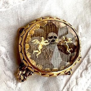 May include: A gold-toned locket with a glass cover, featuring a skull and two winged figures. The locket is decorated with intricate details and has a delicate design.