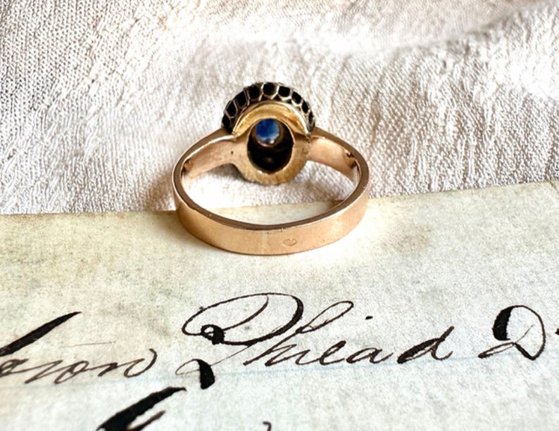 May include: A gold ring with a blue gemstone surrounded by black stones. The ring is on a piece of paper with the words "Philad" written on it.