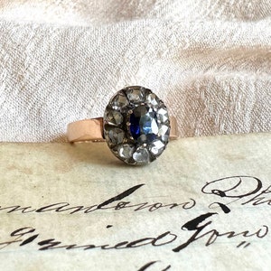 May include: A vintage ring with a blue sapphire surrounded by a halo of small diamonds set in silver. The ring is on a piece of paper with cursive writing.