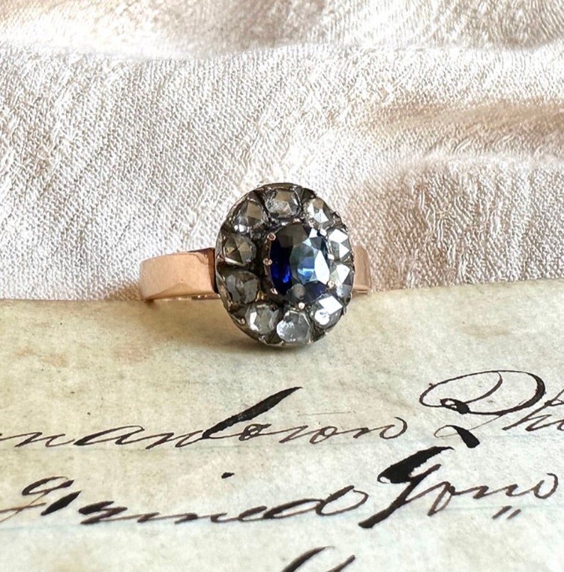 May include: A vintage ring with a blue sapphire surrounded by a halo of small diamonds set in silver. The ring is on a piece of paper with cursive writing.
