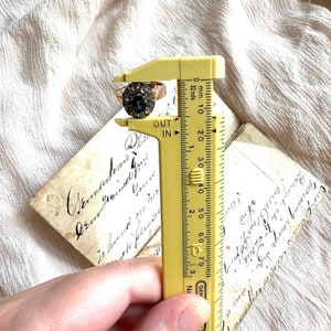 May include: A close-up of a ring with a blue stone surrounded by smaller stones, being measured with a yellow ruler. The ruler shows measurements in inches and millimeters. The ring is approximately 19 millimeters in diameter.