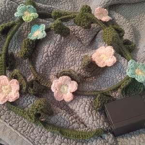Könnte beinhalten: Eine gehäkelte Girlande mit rosa, blauen und grünen Blumen. Die Girlande ist aus grünem Garn gefertigt und mit gehäkelten Blumen verziert. Die Girlande liegt auf einer grauen Stoffoberfläche.