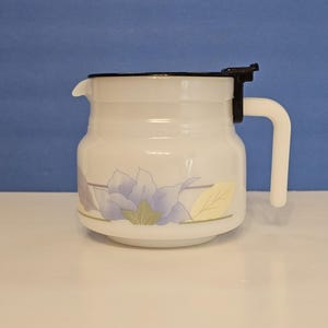 May include: White ceramic pitcher with a black lid and handle. The pitcher features a floral design with purple and yellow accents. The handle is white and the spout is on the left side.