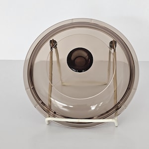 May include: A transparent, brown-tinted glass lid with a dark brown knob. The round lid rests on a white metal stand. The lid is designed for a pot or pan.