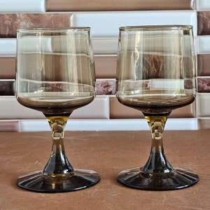 May include: Two clear glass wine glasses with brown tinted bottoms and stems. The glasses have a wide bowl and a round base.