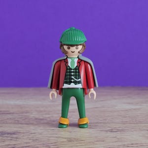 Peut inclure: Une figurine Playmobil de détective, vêtue d'une casquette verte, d'une veste rouge, d'une cape grise, d'un pantalon vert et de chaussures jaunes. La figurine porte une chemise blanche et une cravate verte. Le fond est violet.