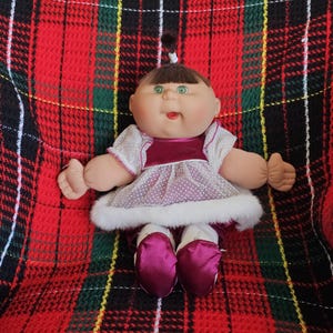 May include: A vintage doll with a round face, green eyes, and a small ponytail. The doll is dressed in a white dress with pink polka dots, a burgundy velvet bodice, and matching shoes. The doll is on a red, black, and green tartan blanket.