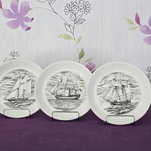 May include: Three white ceramic plates with a nautical theme. Each plate features a detailed black and white illustration of a sailing ship. The plates are displayed on a purple surface.