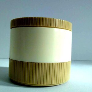 May include: A cylindrical container with a cream-coloured body and ribbed, tan-coloured top and bottom sections. The container is likely made of plastic and has a simple, minimalist design. The background is a light blue.