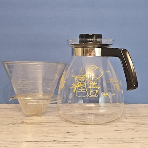 Peut inclure: Une cafetière en verre transparent avec une poignée et un couvercle noirs, et une bande dorée. La cafetière présente des illustrations et du texte jaunes liés à la préparation du café. Un filtre à café en verre transparent et un petit filtre sont à côté.
