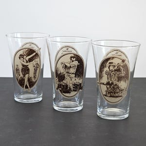 May include: Three clear glass tumblers with brown and white illustrations of athletes. The tumblers are on a dark grey surface.
