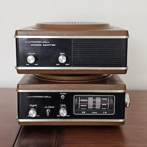 May include: A vintage Wynford Hall stereo adaptor and radio set. The top unit is a stereo adaptor with volume and tone controls. The bottom unit is a radio with AM/FM/MC, volume, and tone controls. Both units have brown wood-grain tops and black faces.