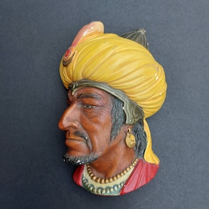 May include: A ceramic wall plaque depicting a man wearing a yellow turban and a red and gold robe. The man has a dark complexion and a serious expression.